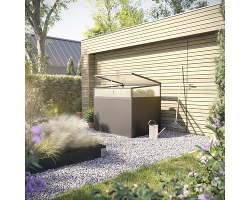 Gartenszene mit einem Pflanztisch aus Metall mit transparenter Abdeckung, einer Gießkanne und einem Rechen vor einer Holzwand.