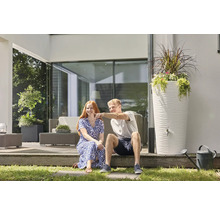 Ein Mann und eine Frau sitzen auf einer modernen Terrasse neben einer hohen weißen Regentonne mit strukturierter Oberfläche und Pflanzschale.
