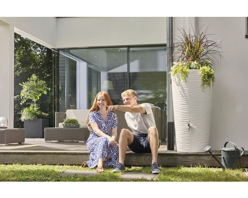 Ein Mann und eine Frau sitzen auf einer modernen Terrasse neben einer hohen weißen Regentonne mit strukturierter Oberfläche und Pflanzschale.