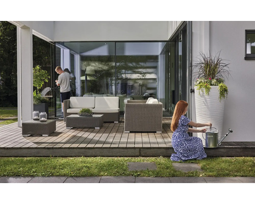 Helle, moderne Holzterrasse mit grauen Loungemöbeln aus Polyrattan, weißen Sitzkissen, Pflanzgefäßen und einer Gießkanne aus Metall.