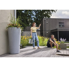 Hohe graue Regentonne in Steinoptik mit integriertem Pflanztopf und Auslaufhahn auf einer Holzterrasse in einem modernen Garten.