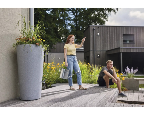 Hohe graue Regentonne in Steinoptik mit integriertem Pflanztopf und Auslaufhahn auf einer Holzterrasse in einem modernen Garten.