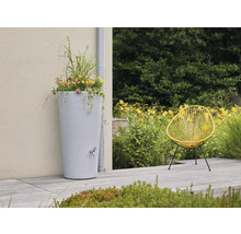 Hohe Regentonne in Steinoptik mit Pflanztopf und Auslaufhahn auf einer hellen Terrasse neben einem Fallrohr, gelber Sessel und Gartenpflanzen.