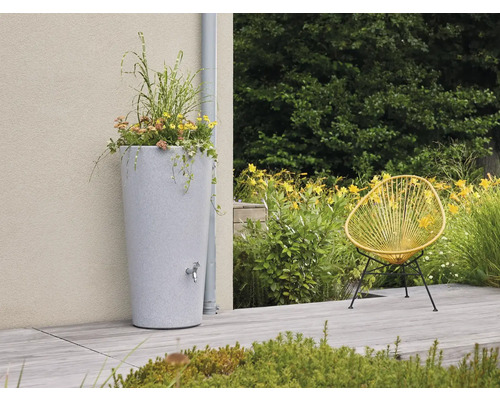 Hohe Regentonne in Steinoptik mit Pflanztopf und Auslaufhahn auf einer hellen Terrasse neben einem Fallrohr, gelber Sessel und Gartenpflanzen.