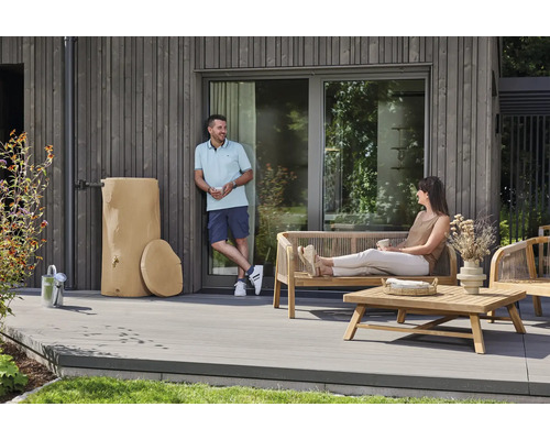 Regenspeicher in Baumstamm-Optik neben Gartenmöbeln aus Holz auf einer modernen Terrasse mit grauen Dielen.