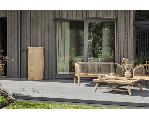 Beige Regentonne an einer grauen Holzfassade auf einer modernen Terrasse mit hölzernen Gartenmöbeln.