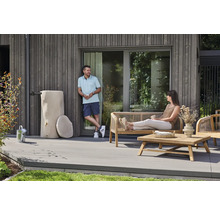 Eine moderne Holzterrasse mit einer dekorativen Regentonne in Baumstammoptik, Gartenmöbeln aus Holz und zwei Personen vor einer Holzfassade.