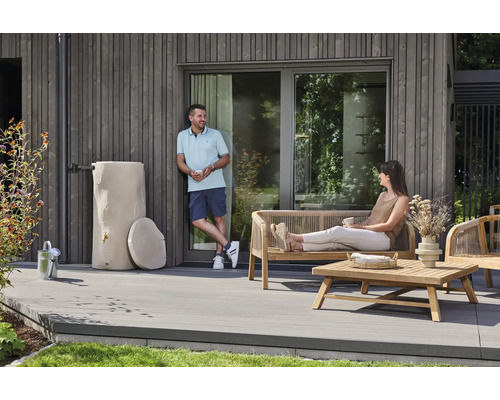Eine moderne Holzterrasse mit einer dekorativen Regentonne in Baumstammoptik, Gartenmöbeln aus Holz und zwei Personen vor einer Holzfassade.