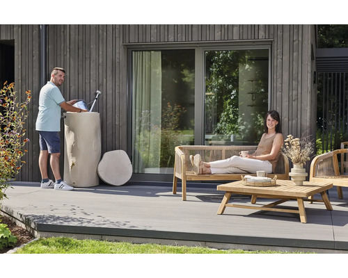 Ein Mann befüllt eine Gießkanne an einer Regentonne in Baumstammoptik auf einer modernen Terrasse mit Gartenmöbeln aus Holz.