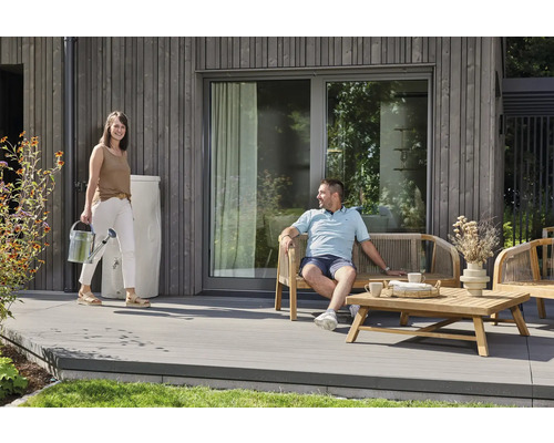 Moderne Holzterrasse mit Gartenmöbeln aus Holz, einer weißen Regentonne an der Hauswand und einer Person mit einer Gießkanne.
