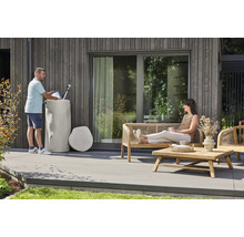 Regentonne in Baumstamm-Optik auf einer Terrasse mit Holzfassade und Gartenmöbeln aus Holz sowie einer sitzenden Frau.