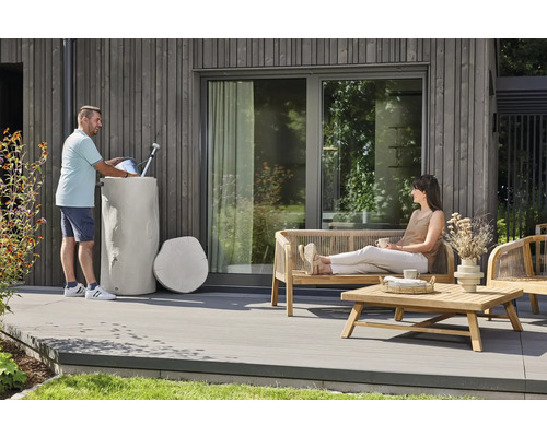 Regentonne in Baumstamm-Optik auf einer Terrasse mit Holzfassade und Gartenmöbeln aus Holz sowie einer sitzenden Frau.