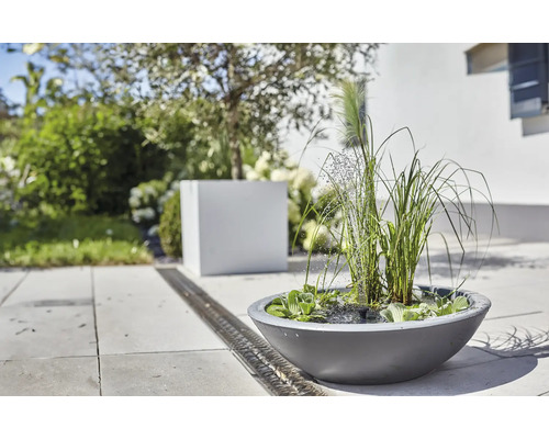 Gartenszene mit einem runden Pflanzgefäß mit Wasserspiel, umgeben von Grünpflanzen und einem Entwässerungsgraben.
