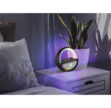 Nachttischszene mit mehrfarbiger Lampe, Lautsprecher und kabelloser Ladestation neben einer Zimmerpflanze