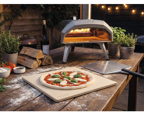 Außenaufnahme eines Ooni Pizzaofens mit Pizza, Pizzaschieber und Zutaten auf einem Holztisch.