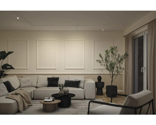 Modernes Wohnzimmer mit großem Ecksofa, dekorativen Wandpaneelen, Schienensystem-Beleuchtung und Zimmerpflanze. Neutrale Farben und warme Beleuchtung.