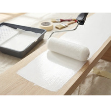 Holzbrett wird mit Farbroller und weißer Farbe gestrichen, Farbwanne, Pinsel und Abdeckband liegen bereit.