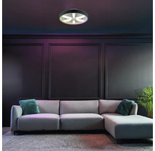 Moderne LED-Deckenleuchte mit Ventilator und RGB-Farbfunktion, montiert in einem dunklen Wohnzimmer mit grauem Sofa und Wandpaneelen.