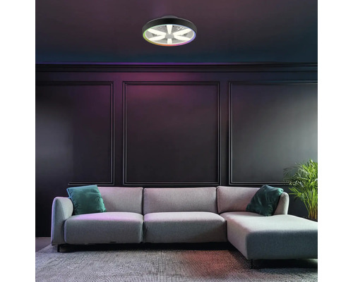 Moderne LED-Deckenleuchte mit Ventilator und RGB-Farbfunktion, montiert in einem dunklen Wohnzimmer mit grauem Sofa und Wandpaneelen.