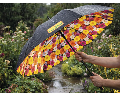 Regenschirm in einem regnerischen Garten mit schwarzer Außenseite, FloraSelf Logo und buntem Blumenmuster auf der Innenseite.