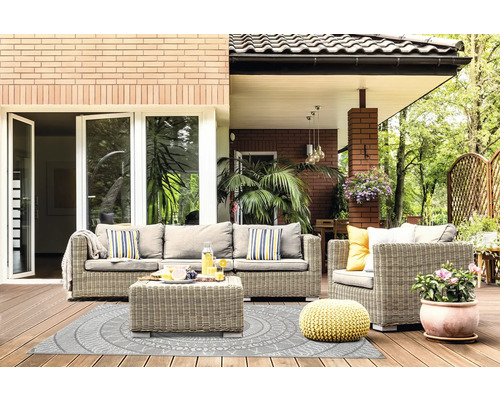 Helle Terrasse mit grauer Rattan-Sitzgruppe, Outdoorteppich mit Kreismuster, gelbem Strick-Pouf und Topfpflanze auf Holzdielen.