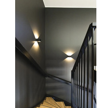Moderne Treppe mit dunklen Wänden, Holzstufen und schwarzem Geländer. Zwei Wandleuchten mit Lichtstrahlen nach oben und unten setzen Akzente.