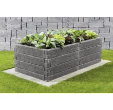 Rechteckiges Hochbeet aus strukturierten grauen Betonsteinen, bepflanzt mit Salat, auf einer gepflasterten Basis im Garten vor einer Steinmauer.