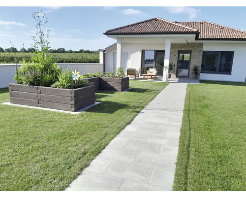 Moderner Garten mit gepflastertem Weg aus hellgrauen Platten, zwei Hochbeeten aus dunklen Modulsteinen und einem Bungalow im Hintergrund.