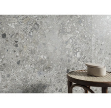 Wandfliesen in Terrazzo-Optik mit grauen und weißen Steineinschlüssen. Im Vordergrund ein runder Hocker mit Geflechtsitz und eine Keramikdose.