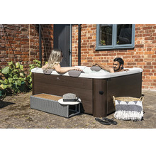 Quadratischer Whirlpool in Holzoptik mit Außenverkleidung, zwei Personen entspannen im Wasser auf einer gepflasterten Terrasse vor einer Ziegelwand.
