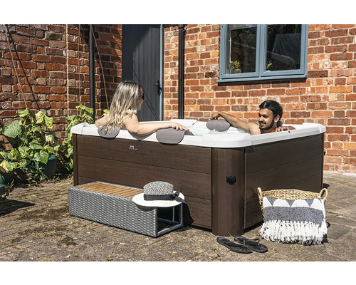 Quadratischer Whirlpool in Holzoptik mit Außenverkleidung, zwei Personen entspannen im Wasser auf einer gepflasterten Terrasse vor einer Ziegelwand.