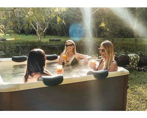 Drei Frauen entspannen in einem rechteckigen Außen-Whirlpool mit Kopfstützen in einem Garten an einem Teich.