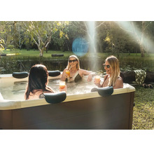 Drei Frauen entspannen in einem Whirlpool im Garten neben einem Teich. Der Whirlpool hat eine dunkle Verkleidung und Kopfstützen.