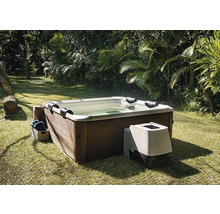 Quadratischer Whirlpool mit Außenverkleidung in Holzoptik und externer Steuereinheit in einem hellen, weitläufigen Garten mit Palmen.