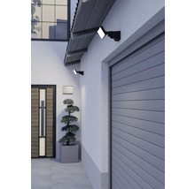 Zwei schwarze LED-Außenstrahler montiert an einer Hauswand über einem grauen Garagentor und neben einer Holztür in einem modernen Eingangsbereich.