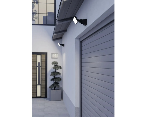Zwei schwarze LED-Außenstrahler montiert an einer Hauswand über einem grauen Garagentor und neben einer Holztür in einem modernen Eingangsbereich.