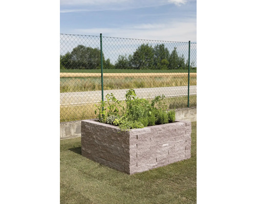 Hochbeet aus Steinblöcken im Garten, bepflanzt mit Kräutern und Pflanzen vor einem Zaun.