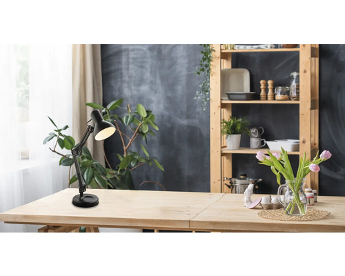 Schwarze, verstellbare Schreibtischleuchte auf einem hellen Holztisch in einem hellen Zimmer mit Zimmerpflanzen und einem Holzregal.