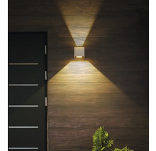 Quadratische weiße Außenwandleuchte an einer Holzwand neben einer dunklen Tür, die warmes Licht nach oben und unten wirft.