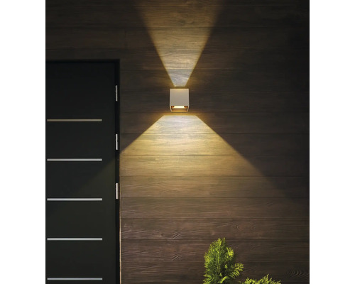 Quadratische weiße Außenwandleuchte an einer Holzwand neben einer dunklen Tür, die warmes Licht nach oben und unten wirft.