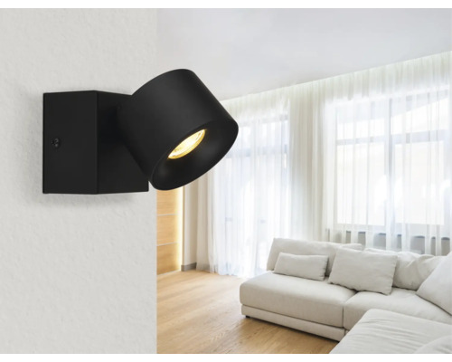 Moderner schwarzer verstellbarer Wandstrahler in einem hellen, geräumigen Wohnzimmer mit weißem Sofa und transparenten Vorhängen.