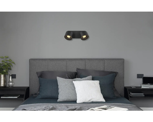 Moderne schwarze Wandleuchte mit zwei Spots über einem grauen Bett in einem Schlafzimmer.