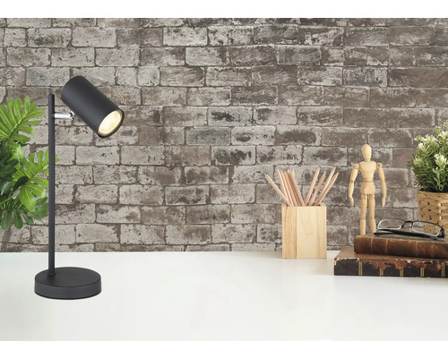 Schwarze Schreibtischlampe auf weißem Tisch vor einer Backsteinwand, neben Stiftehalter, Gliederpuppe und Büchern.