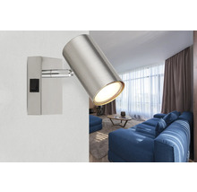 Moderner silberner Wandstrahler aus Metall mit zylindrischem Kopf und Schalter an einer Wand in einem hellen Wohnzimmer mit blauem Sofa.