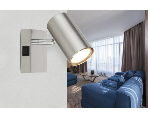 Moderner silberner Wandstrahler aus Metall mit zylindrischem Kopf und Schalter an einer Wand in einem hellen Wohnzimmer mit blauem Sofa.