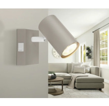 Verstellbarer Wandstrahler in einem hellen Wohnzimmer mit Sofa und Couchtisch. Die Lampe hat einen zylindrischen Schirm und einen Wippschalter.