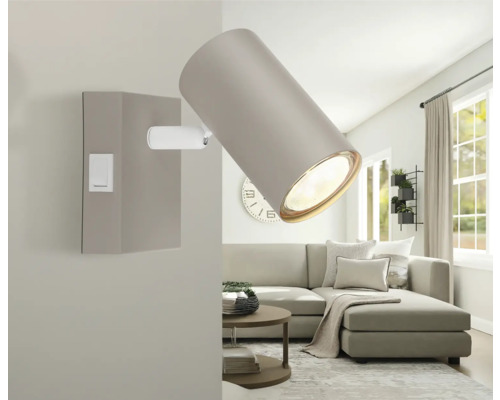 Verstellbarer Wandstrahler in einem hellen Wohnzimmer mit Sofa und Couchtisch. Die Lampe hat einen zylindrischen Schirm und einen Wippschalter.