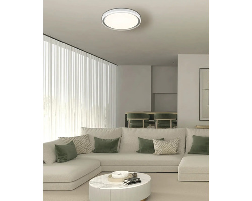 Modernes Wohnzimmer mit runder LED-Deckenleuchte, großem weißem Ecksofa mit grünen Kissen und einem runden Couchtisch aus Marmor.