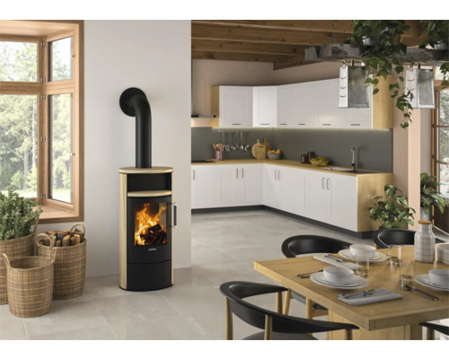 Moderner Kaminofen mit heller Steinverkleidung und schwarzem Ofenrohr in einer hellen Küche mit weißer Küchenzeile und Holztisch.