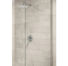 Moderne Dusche mit verchromtem Regenduschkopf, Einhebelmischer und hellgrauen Wandfliesen in Steinoptik hinter einer Glaswand.